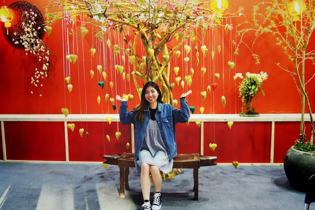Red wall background with golden hearts tree at a flower festival, Amy sitting on a bench in front of the tree